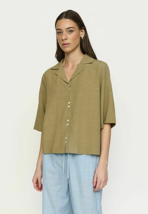 Vrouw draagt een loszittend olijfgroen overhemd met knopen en halve mouwen, en lichtblauwe broek met trekkoord, staand tegen een effen achtergrond.