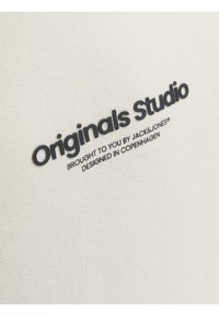 Cremefarvet bomuldsstof med fremtrædende sort tekst, der lyder "Originals Studio" og "Brought to you by JACK&JONES, Designed in Copenhagen."