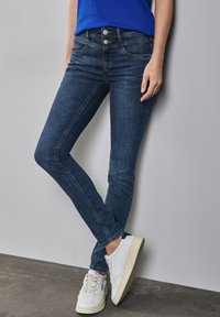 Jean skinny bleu foncé à taille haute, avec cinq poches et deux fermetures à bouton, associés à des baskets blanches sur un fond gris.