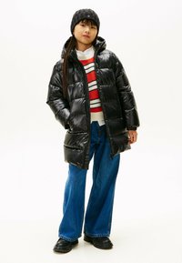 Doudoune noire avec une finition brillante, coupe oversize, fermeture éclair à l'avant et capuche, associée à un pull rayé rouge et à un jean bleu évasé.