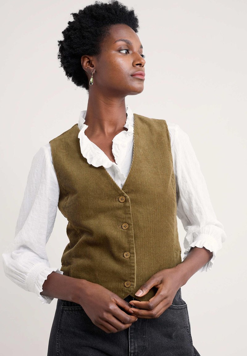 Maslinasto zelena kordura prsluk s pet drvenih dugmadi, uparen s bijelom bluzom s volanima. Model pozira s rukama sklopljenim na struku.