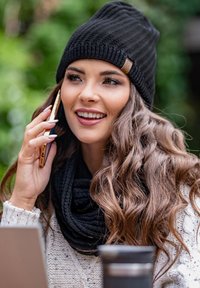 Gorro de punto negro con una etiqueta de cuero, combinado con una bufanda de punto negra. La persona tiene el cabello largo y ondulado de color marrón y sostiene un teléfono inteligente.