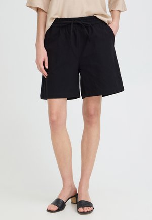 Person trägt schwarze kniehohe Shorts mit Kordelzug, beiges Oberteil und schwarze offene Sandaletten mit Absatz, steht mit Händen in den Taschen.