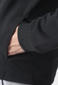 Mano insertada en el bolsillo lateral de una chaqueta negra con puños acanalados, mostrando parte del antebrazo y la muñeca.