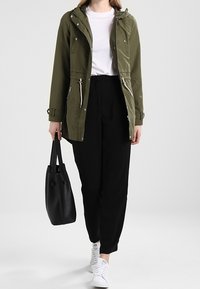 Nainen, joka pukeutunut oliivinvihreään takkiin, valkoiseen t-paitaan, mustiin housuihin, valkoisiin lenkkareihin, kantaa mustaa kangaskassia ja kävelee eteenpäin valkoista taustaa vastaan.