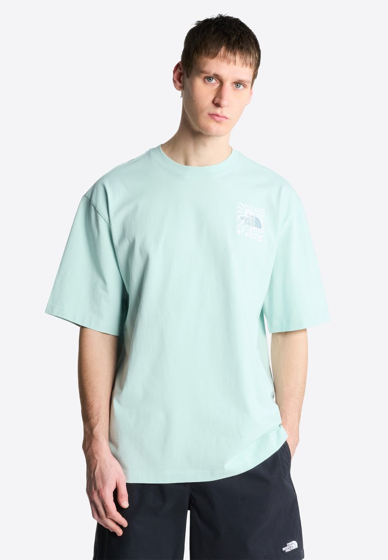 Junger Mann steht, trägt ein hellblaues The North Face T-Shirt und schwarze Shorts, die Hände entspannt an den Seiten, nach vorne blickend vor einem schlichten weißen Hintergrund.