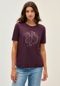 Camiseta marrón con mangas cortas, cuello redondo, que presenta un gráfico circular en tonos más claros y texto. Tejido de algodón suave, corte relajado.