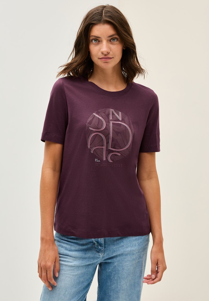 Camiseta marrón con mangas cortas, cuello redondo, que presenta un gráfico circular en tonos más claros y texto. Tejido de algodón suave, corte relajado.