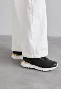 Baskets en cuir noir avec un accent doré sur le talon, semelle blanche et finition texturée, associées à un pantalon blanc à jambes larges.