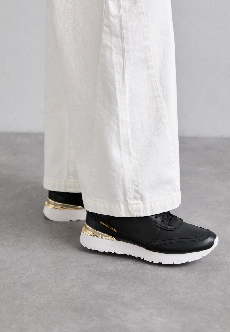 Baskets en cuir noir avec un accent doré sur le talon, semelle blanche et finition texturée, associées à un pantalon blanc à jambes larges.