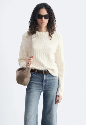 Femme aux cheveux bouclés portant des lunettes de soleil noires, un pull en maille crème, un jean bleu, une ceinture marron, et un sac à bandoulière en daim taupe.