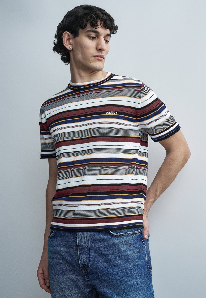 Young man with curly dark hair wears multicolored horizontal striped Missoni T-shirt and blue jeans, looking to his left with hand on hip.