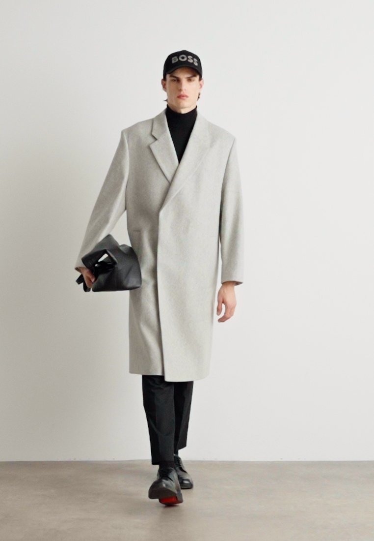 Manteau en laine gris avec col châle, porté sur un pull noir à col roulé. Le mannequin tient un sac à main noir et porte une casquette noire. Pantalons fins noirs et chaussures.