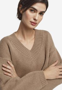 Pull en maille beige doux avec un col en V, des manches surdimensionnées et des détails côtelés. Le tissu présente une finition texturée et confortable.
