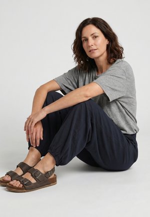 Frau mit dunkel welligem Haar, die ein graues T-Shirt, marineblaue Jogginghosen und braune Sandalen mit Schnallen trägt, sitzt mit überkreuzten Beinen auf hellem Hintergrund.