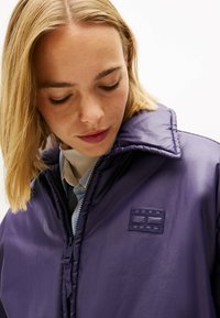 Giacca in pelle viola con colletto alto, caratterizzata da un distintivo testurizzato e chiusura con zip. Sotto, è visibile una camicia azzurro chiaro con colletto.