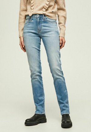 Henkilö pukeutuneena vaaleansinisiin slim-fit farkkuihin, beigeen neulepuseroon ja mustiin paksupohjaisiin nilkkureihin yksivärisellä taustalla.