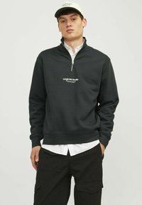 Sudadera gris oscura con cuello con cremallera, que presenta un logo blanco en el pecho. Se lleva sobre una camisa blanca y pantalones cargo negros.
