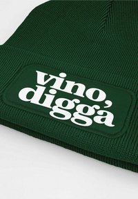 Green knit beanie with ribbed texture, featuring a rectangular white patch that reads "vino', diggà" in bold letters.