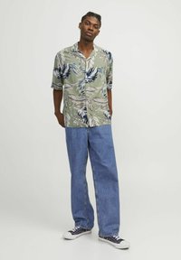 Camisa de manga corta verde con estampado floral y patrones de hojas azules y beige, combinada con jeans holgados de color azul claro y zapatillas de deporte marinas.