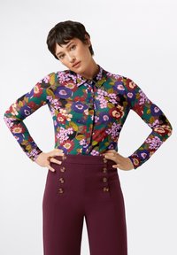 Blusa floreale con maniche lunghe, caratterizzata da uno sfondo verde e fiori di vari colori. Abbinata a pantaloni a vita alta di un profondo borgogna con dettagli di bottoni.
