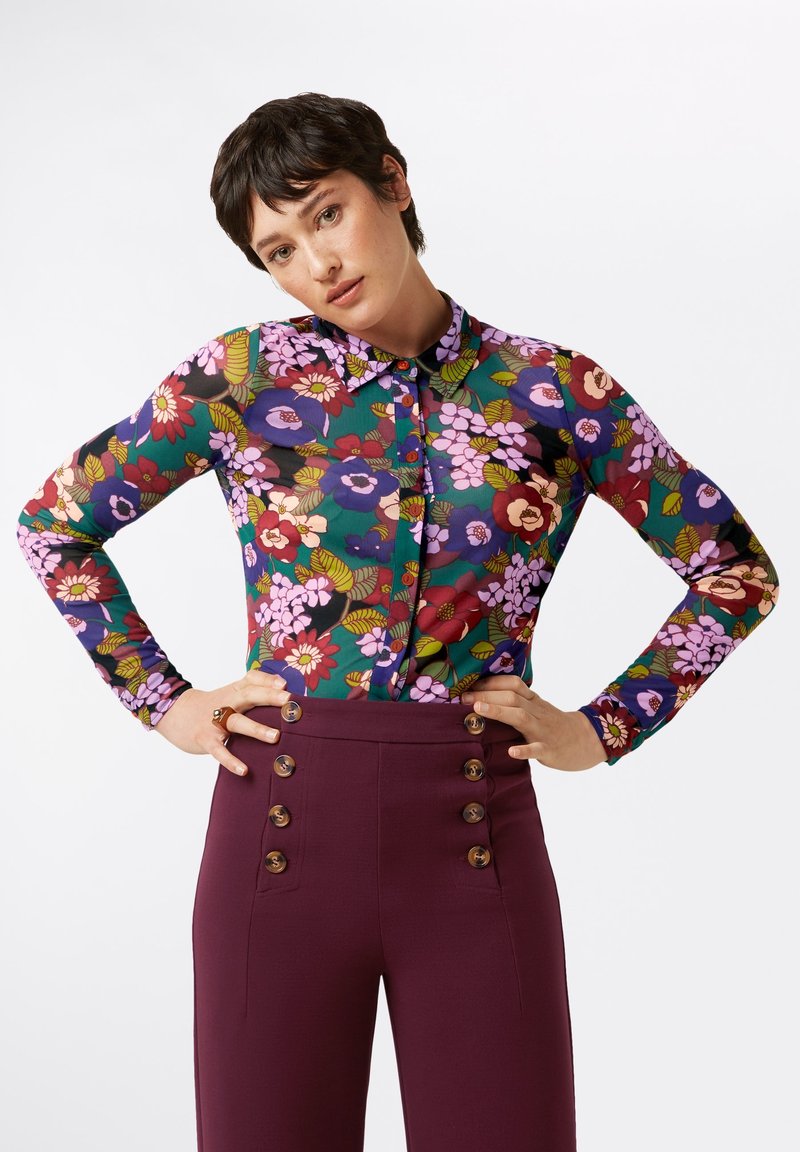 Blusa floreale con maniche lunghe, caratterizzata da uno sfondo verde e fiori di vari colori. Abbinata a pantaloni a vita alta di un profondo borgogna con dettagli di bottoni.