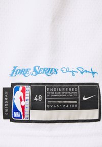 Maglia bianca con texture a rete. Presenta il testo blu "Lore Series" e la firma di Elgin Baylor. Etichetta nera con logo NBA, taglia 48 e marchio Nike.