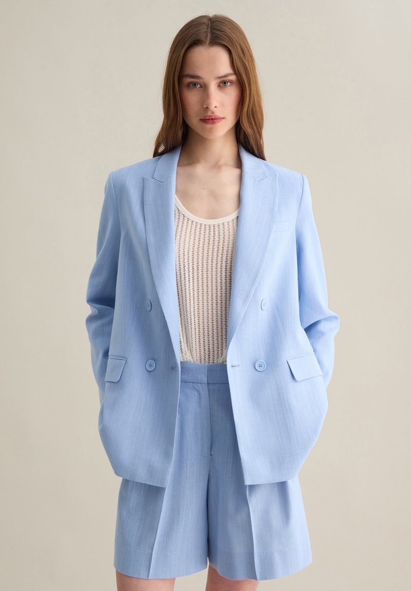 Blazer su misura di un blu chiaro con bottoni doppi, abbinato a pantaloni corti coordinati e a un top a maglia color crema. Tessuto morbido e vestibilità rilassata.