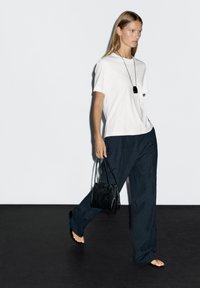 Maglietta bianca, pantaloni larghi blu scuro con una sottile trama e una piccola borsa nera. La modella è scalza, su uno sfondo neutro.