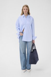 Mujer que lleva una camisa azul claro de botones grande, vaqueros de pierna ancha, zapatos blancos puntiagudos y sostiene un gran bolso tote azul marino.
