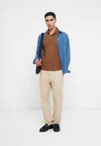 Brūns polo krekls zem džinsa krekla, bēšas bikses, melni apavi un soma. Gluda auduma, brīvas formas un neitrālu krāsu palete.