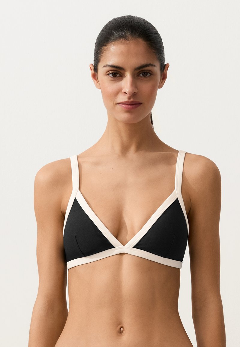 Femme aux cheveux foncés portant un soutien-gorge noir triangulaire avec bordure blanche, debout devant un fond clair uni, face à l'avant.