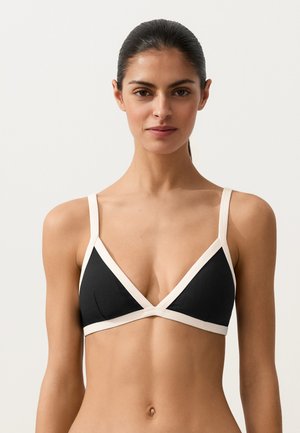 Žena s tmavými vlasy v černé trojúhelníkové podprsence s bílým lemem, stojící na jednoduchém světlém pozadí, čelem vpřed.