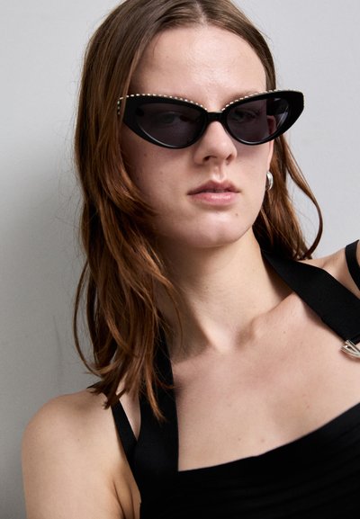 Lunettes de soleil noires en forme d'œil de chat avec des verres foncés et des accents en strass le long du bord supérieur, sur un fond neutre.