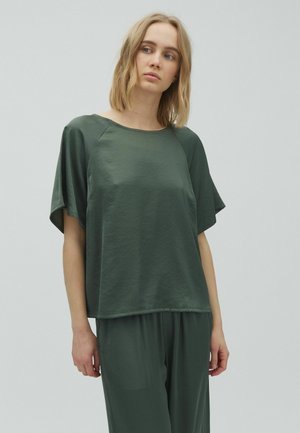 Woman with shoulder-length blonde hair wearing loose dark green short-sleeve top and matching pants, standing against light gray background.