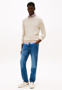 Pull en tricot beige sur une chemise à col rayée, associé à un jean bleu et des baskets blanches. Le fond uni met en valeur la tenue.