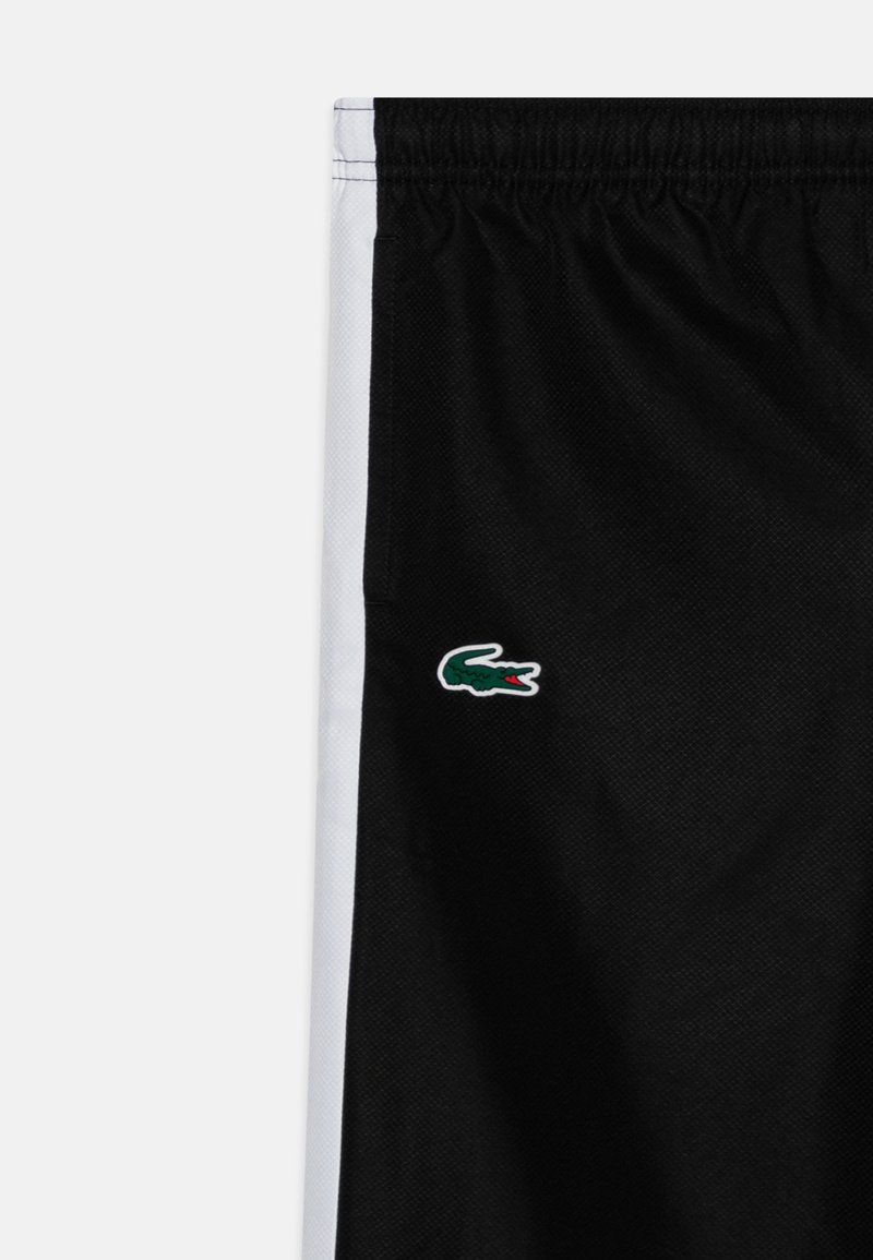 Shorts de sport noirs avec des rayures latérales blanches, arborant un logo de crocodile vert en bas à gauche. Fabriqués en tissu en maille respirant.