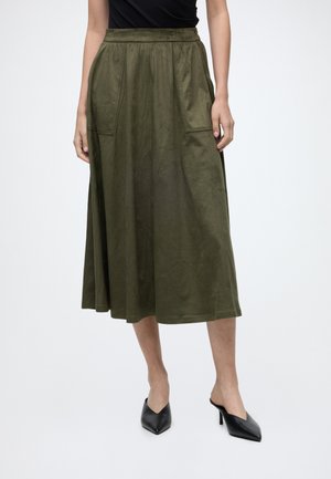 Femme portant une jupe midi vert olive avec de grandes poches avant et des mules à talons noires à bout pointu, debout devant un fond uni.