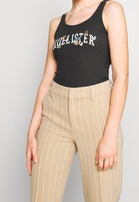 Svart ribbad linne med "HOLLISTER" grafik, matchad med beige randiga högmidjade byxor. Noterbara detaljer inkluderar axelband.