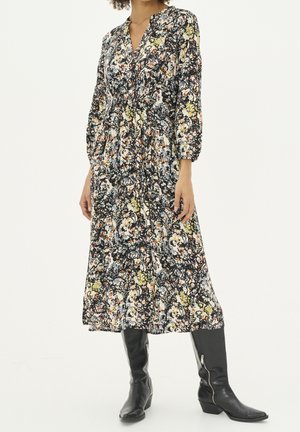 Kvinde iført en lang blomsterkjole med trekvartærmer, kombineret med sorte læderstøvler, der går til midt på læggen, stående foran en ensfarvet baggrund.