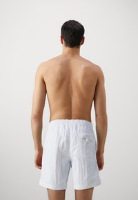 Pantaloni scurți bărbați albi, texturati, cu un brâu elastic și un buzunar pe spate, prezentati din spate pe un fundal deschis.