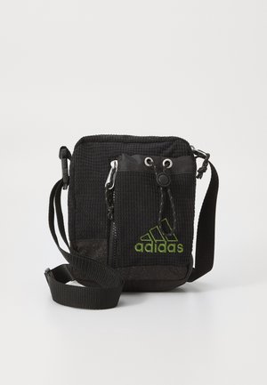 Petit sac bandoulière noir Adidas avec logo vert, poche avant zippée, fermeture à cordon et bandoulière ajustable sur fond blanc.