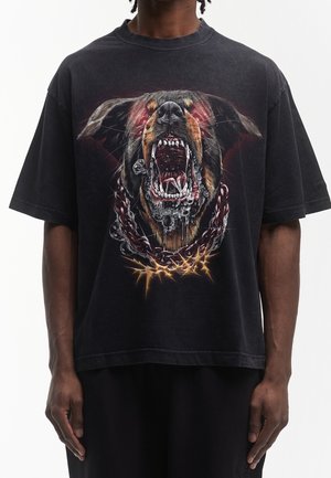 Personne portant un T-shirt noir avec un dessin d’un chien grognant, gueule ouverte, collier en chaîne métallique et fil barbelé en bas.