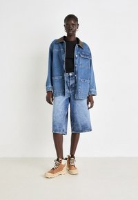 Veste en jean avec col marron, quatre poches avant, associée à un short en jean bleu clair jusqu'aux genoux et des bottes de randonnée beiges avec des lacets noirs.