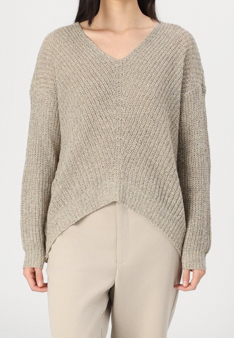 Femme portant un pull en maille beige texturée avec un col en V et un ourlet asymétrique, associé à un pantalon beige clair.