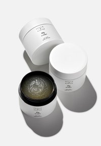Contenitori cilindrici bianchi con coperchi neri, etichettati "Pestle & Mortar Nimbu Body Scrub." Un barattolo è aperto, rivelando uno scrub di colore verde chiaro all'interno.