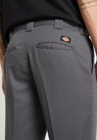 Šedé kalhoty vyrobené z odolné tkaniny, s jednou zadní kapsou a černou nášivkou s logem Dickies a poutky na pás. Hladká textura a přizpůsobený střih.
