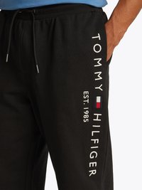Pantaloni da tuta neri in cotone, con ricami bianchi riportanti il marchio "TOMMY HILFIGER" e "EST. 1985" lungo la gamba sinistra. Vita con cordino.