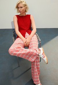 Röd ärmlös topp med texturerad tyg, knapppdetaljer; rosa rutig vida byxor; vita sneakers med röda accenter; sittande på en silverfärgad stol.