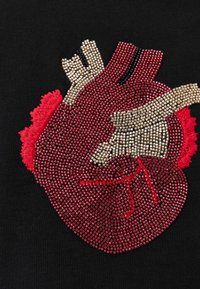 Αναλυτική ανατομική καρδιά φτιαγμένη από μικρές κόκκινες, χρυσές και μαύρες χάντρες σε μαύρο ύφασμα, που δείχνει φλέβες και αρτηρίες.